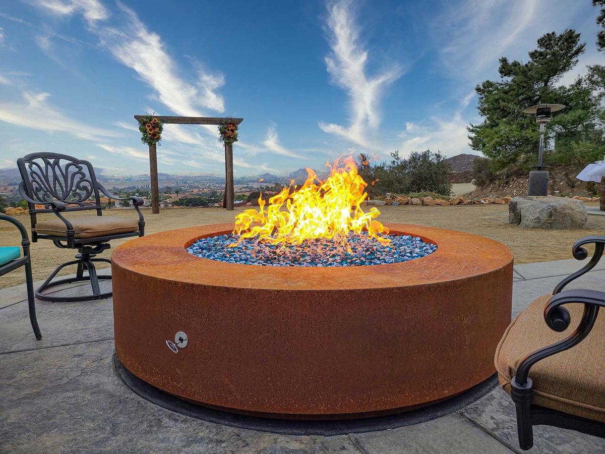 Outdoor Plus Unity gas fire pit in aged Corten steel in an outdoor gathering area — manufacturer photograph